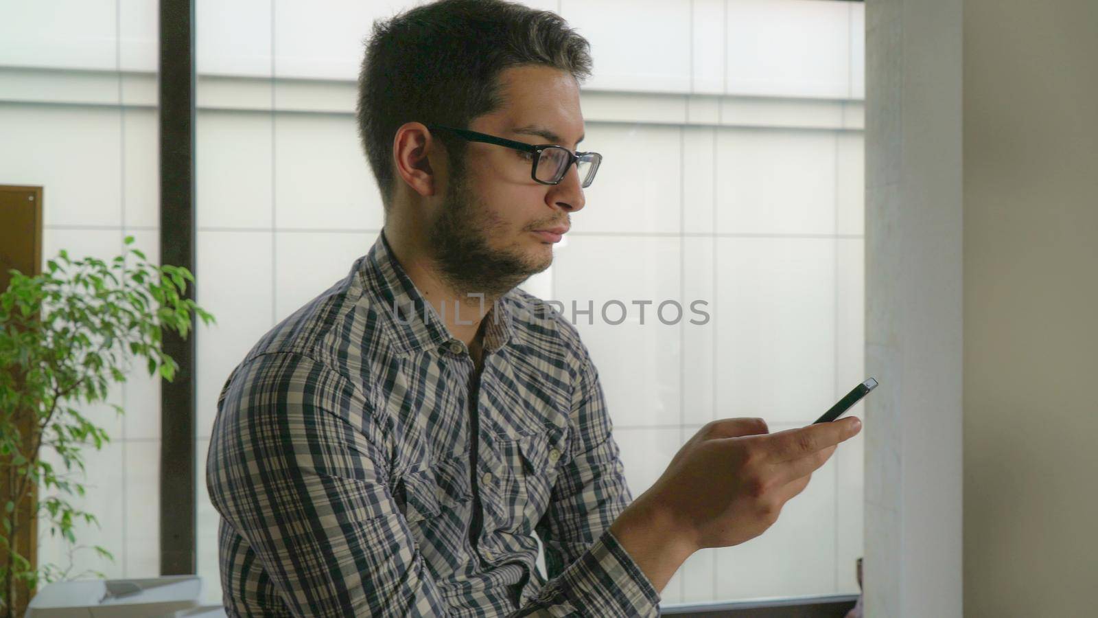 Successful guy in own office with big windows writing sms on cell. Smart businessman with glasses wearing in casual checkered shirt using app on telephone and chatting in social networking service.