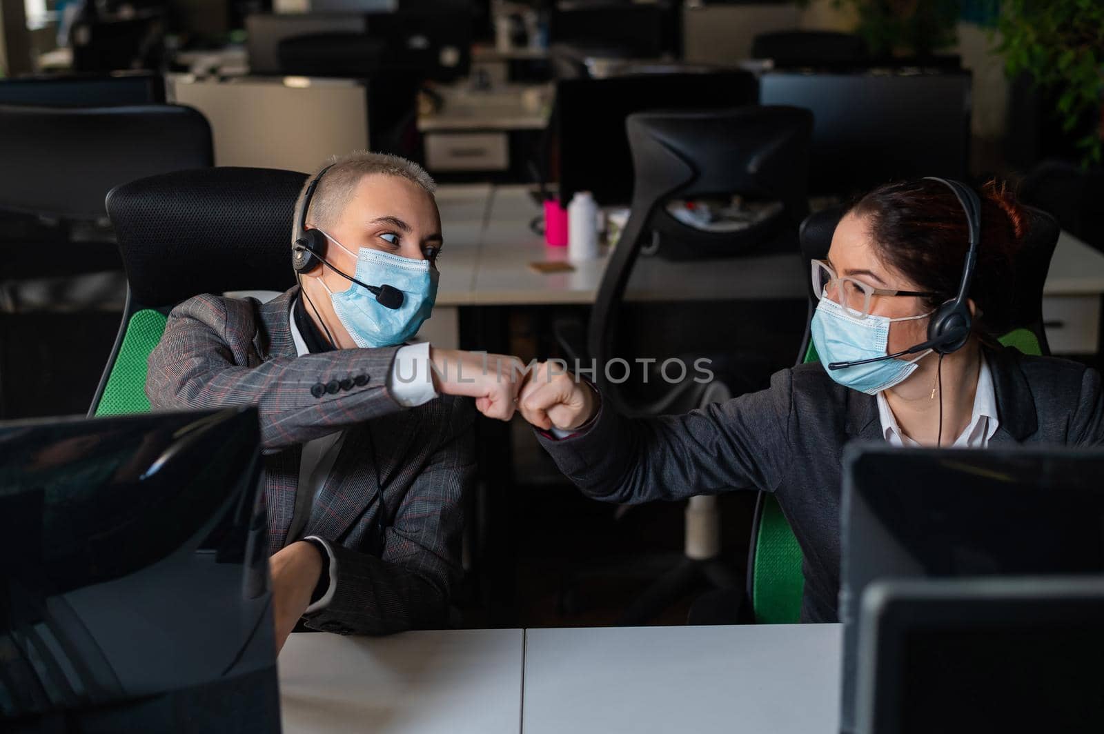 Two business women in masks are giving a high five while sitting at one desk in the office.