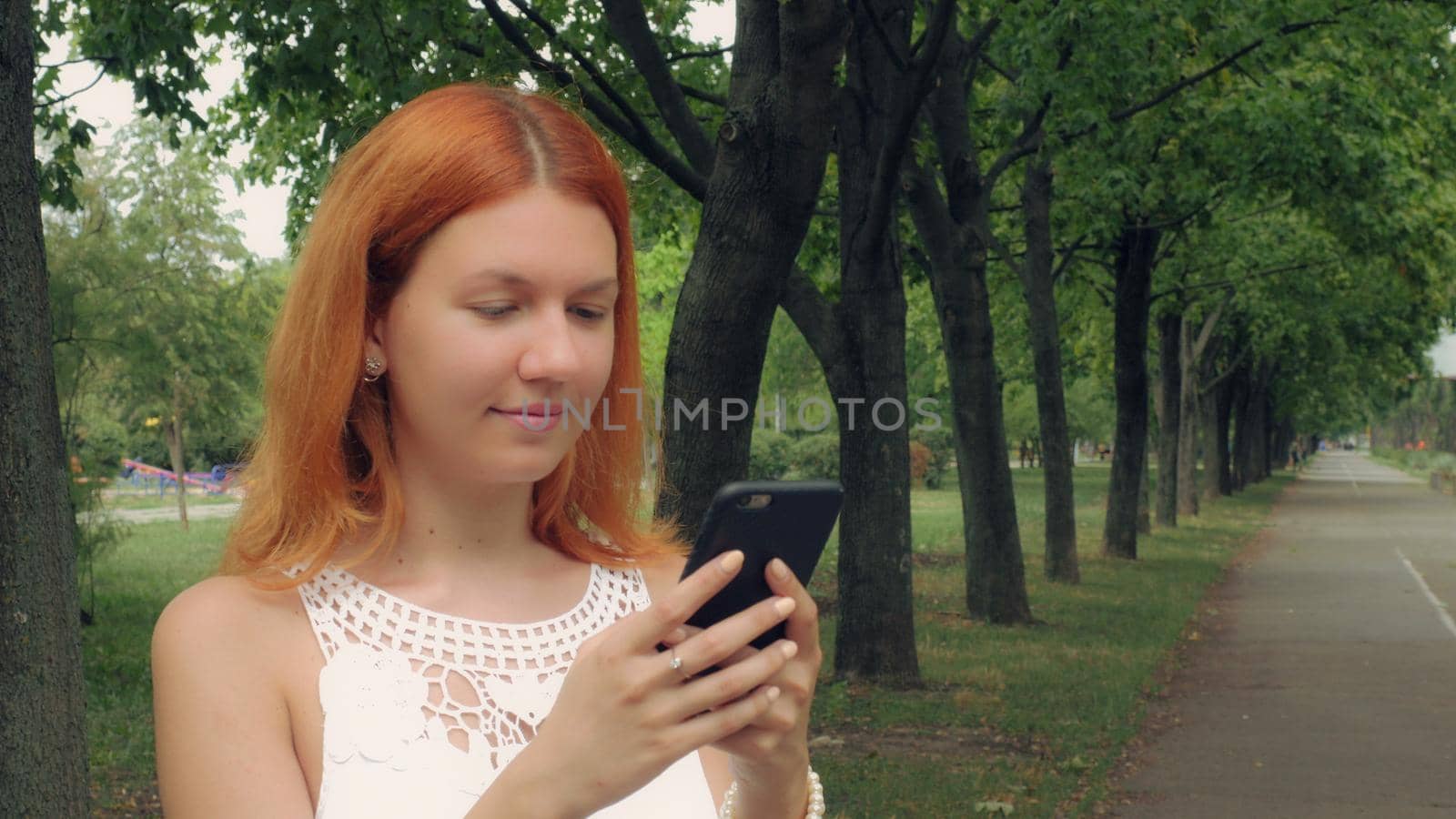 Redheaded woman texting on mobile phone in city with green nature summer season outdoors. Beautiful millennial girl using internet send email or sharing in social media or typing message on touch screen smartphone.