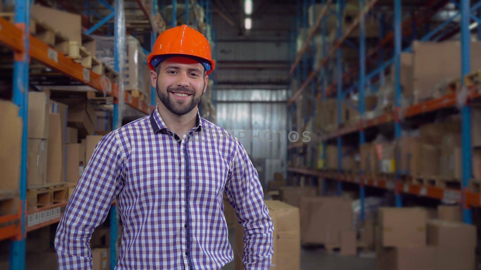 smiling manager near metal racks with boxes wearing casual shirt and orange hard hat. Portrait worker posing in warehouse at work. Happy friendly adult men with beard looking at the camera with smile.