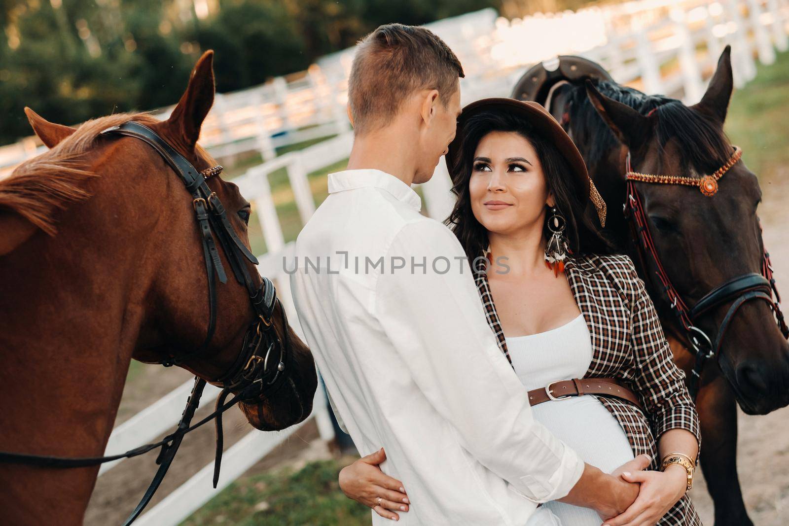 a pregnant girl in a hat and her husband in white clothes stand next to the horses near the horse corral.Stylish pregnant woman with a man with horses.Family.