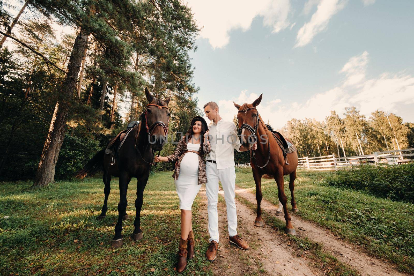 a pregnant girl in a hat and her husband in white clothes stand next to horses in the forest in nature.Stylish pregnant woman with a man with horses.Family