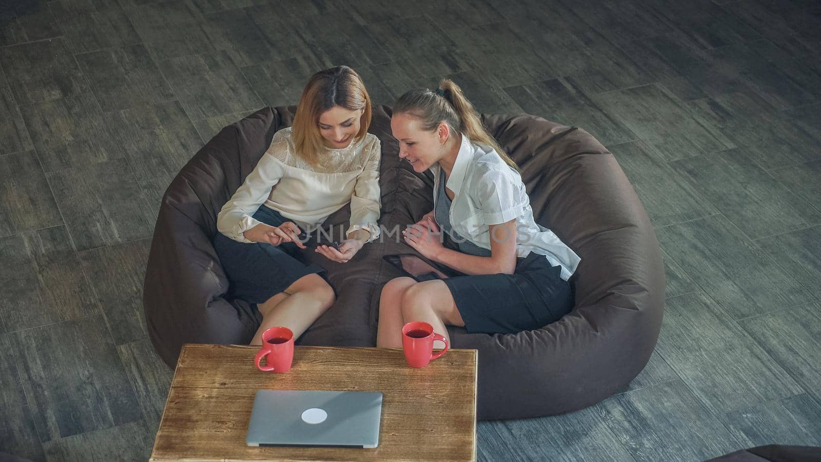 Two attractive business woman relaxing on a sofa and watching photo on the phone. Smile and discuss photos, pleasing appearance. Static video