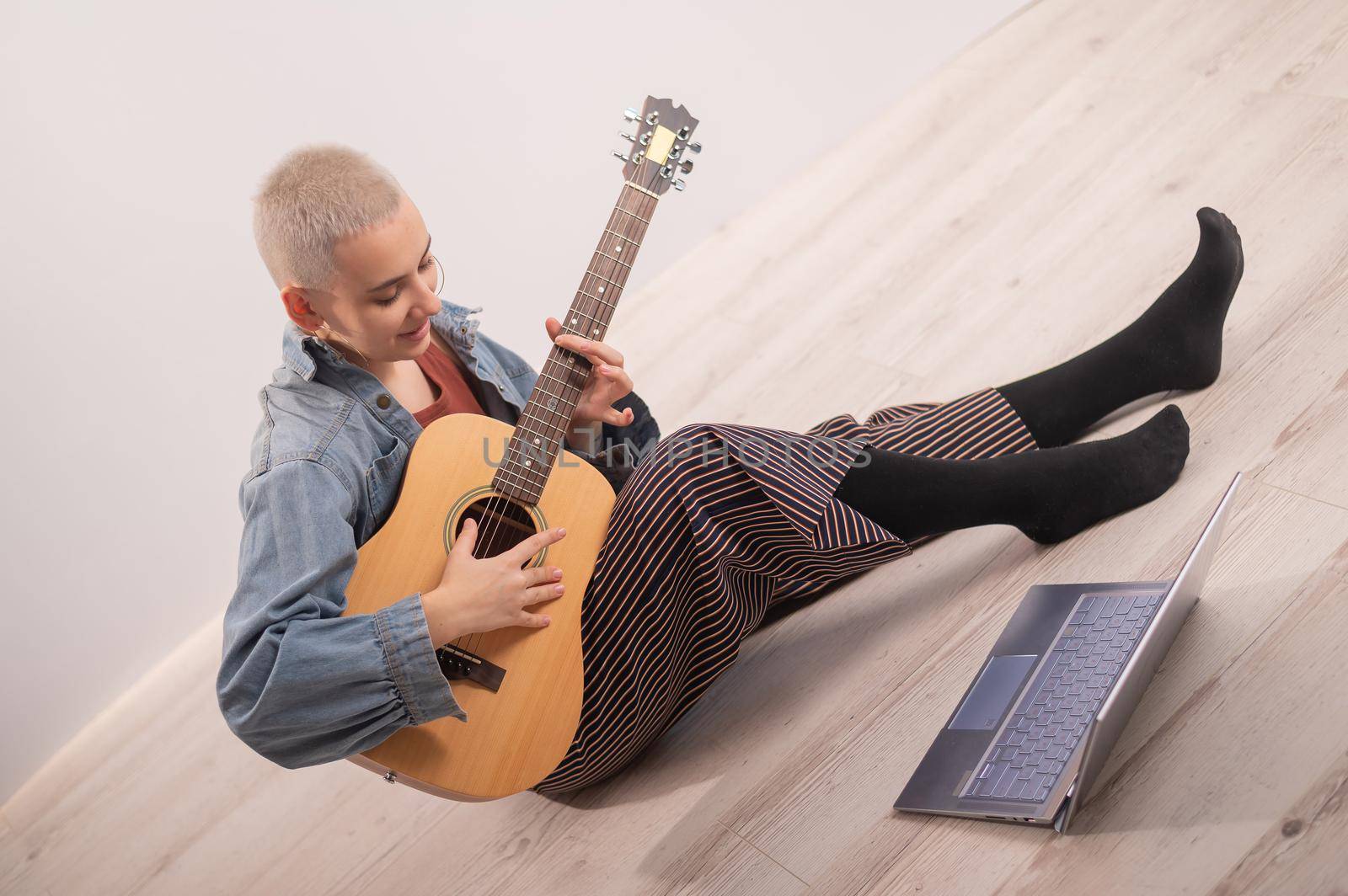 Young hipster woman sitting on the floor at home and watching guitar tutorials on laptop. Distance learning music in quarantine.