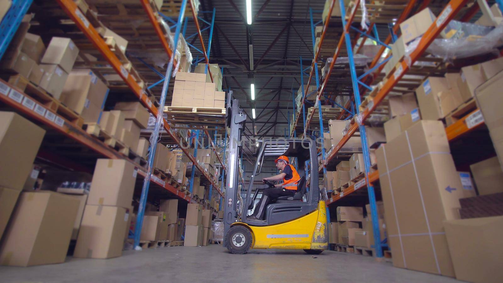 Worker in warehouse riding on forklift trucks with cardboard boxes. Man wearing in uniform hard hat and orange safety vest transporting goods on metal racks.