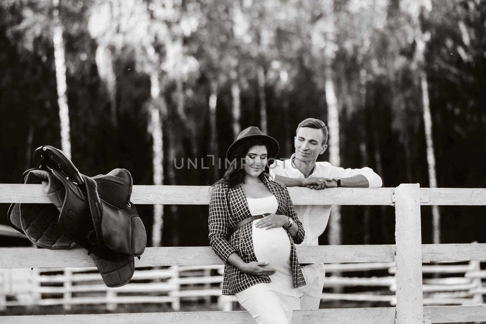 a pregnant girl in a hat and her husband in white clothes stand next to a horse corral at sunset.a stylish couple is waiting for a child in nature. black and white photo by Lobachad