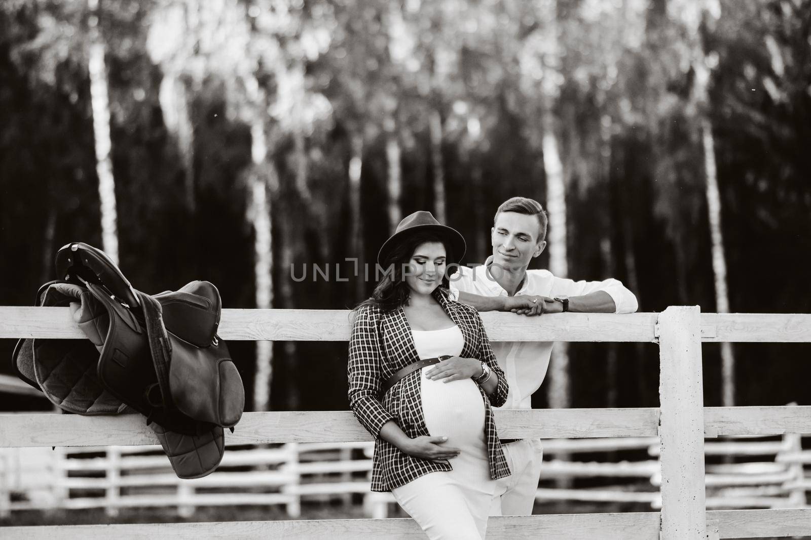 a pregnant girl in a hat and her husband in white clothes stand next to a horse corral at sunset.a stylish couple is waiting for a child in nature. black and white photo.