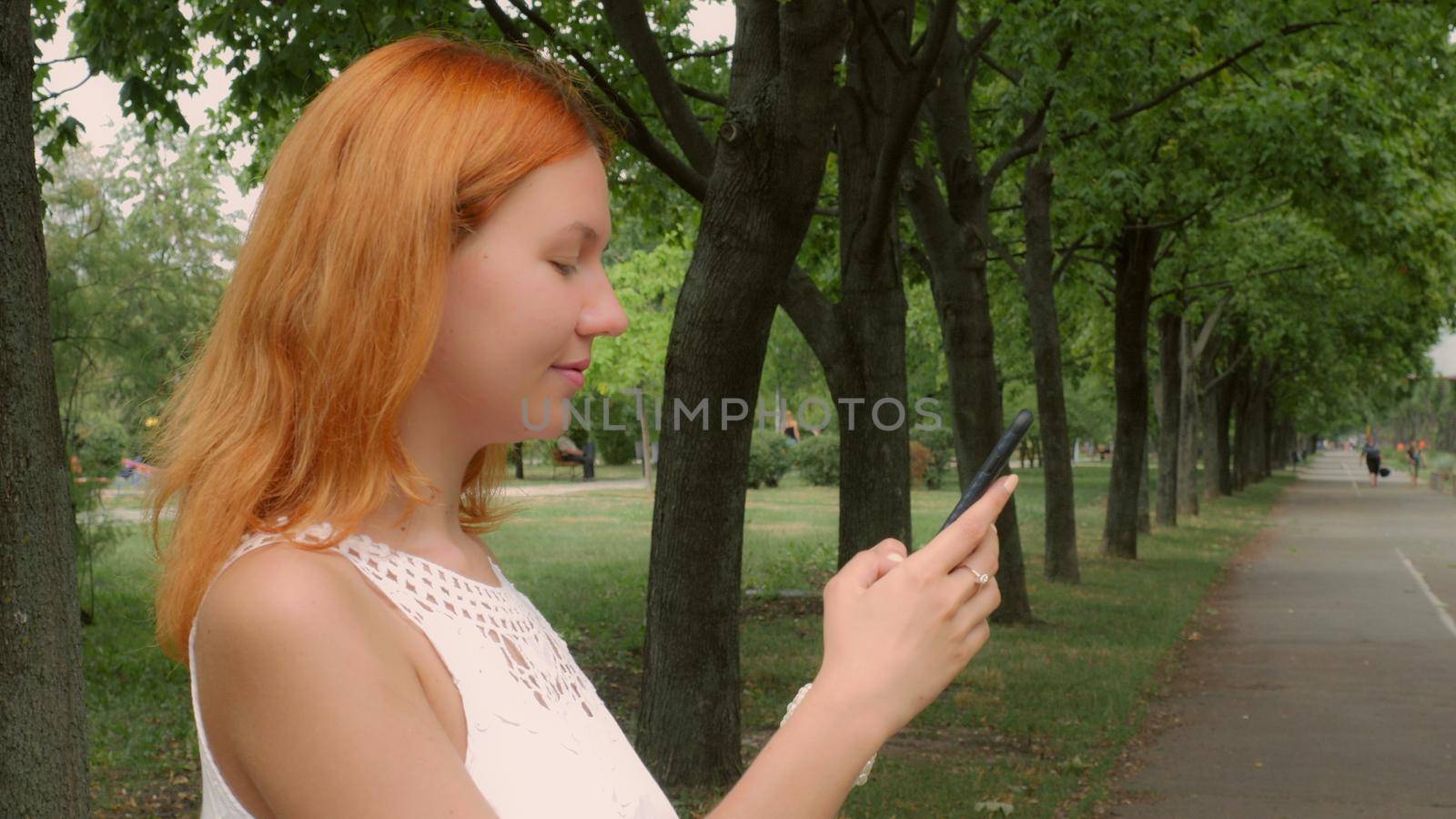Young ginger student standing on the street in park or city using smart phone messaging. Beautiful millennial girl using wifi send email or sharing in social media or typing message on touch screen smartphone.