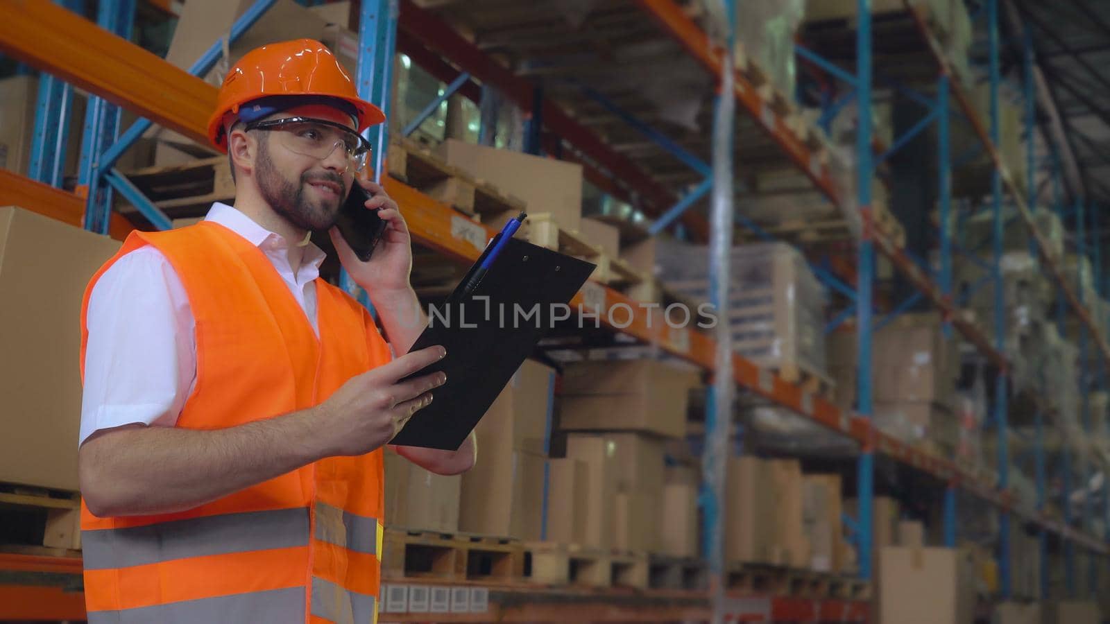 Portrait manager at work in warehouse has phone conversation with client. Handsome worker talking by smartphone discussing the logistics. Man wearing hard hat and orange vest