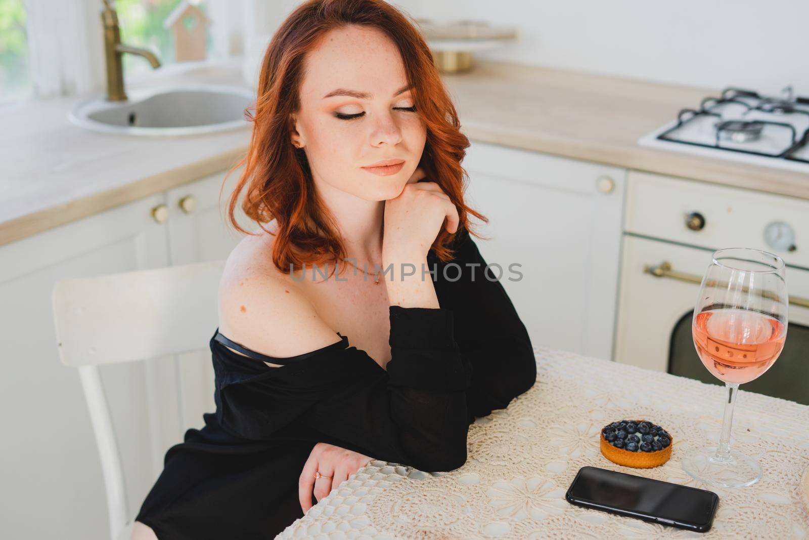 A beautiful girl with red hair holding a smartphone. There's a glass of rose wine on the table. The girl is resting.