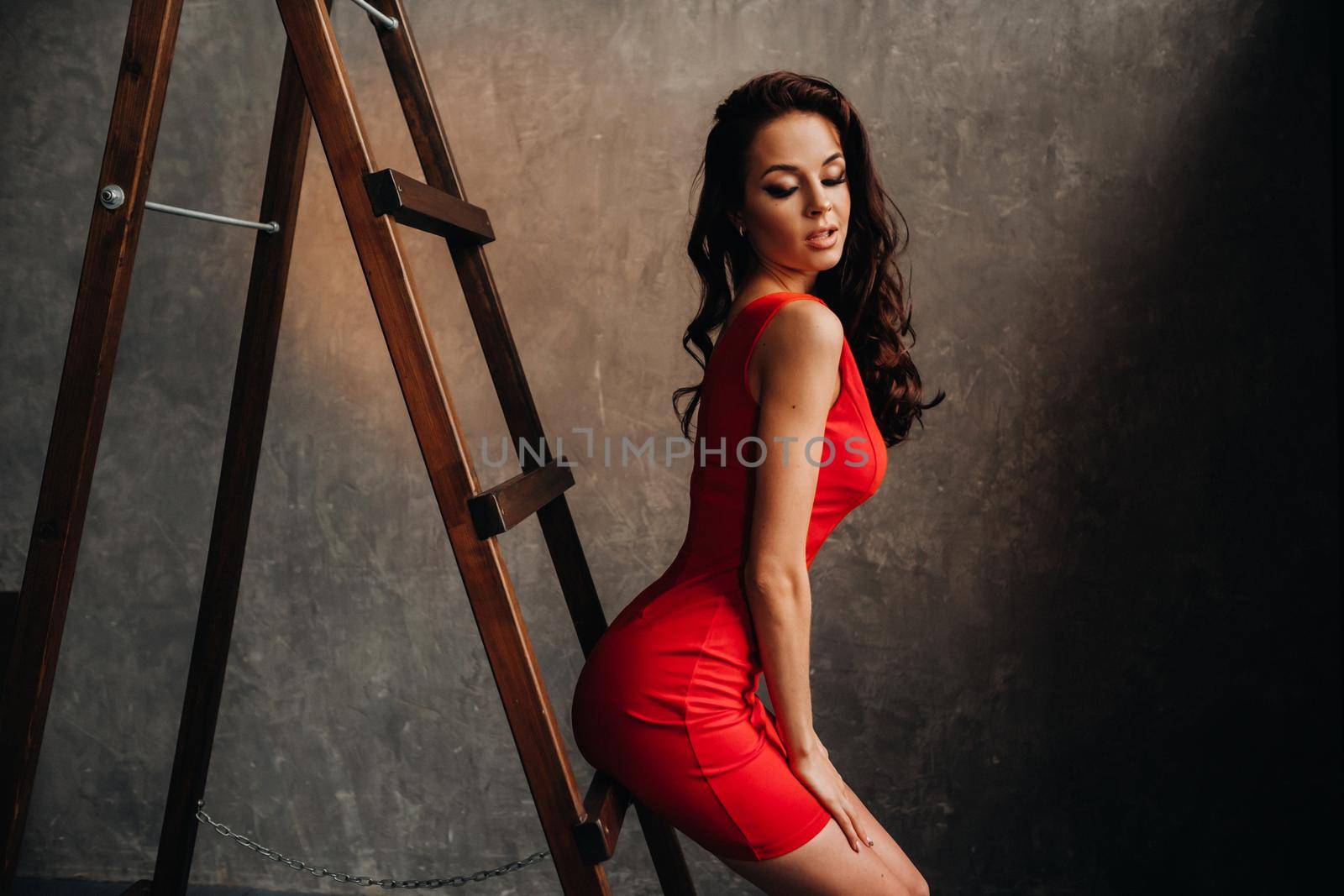 a brunette with long hair in a red dress poses in the studio near the stairs.