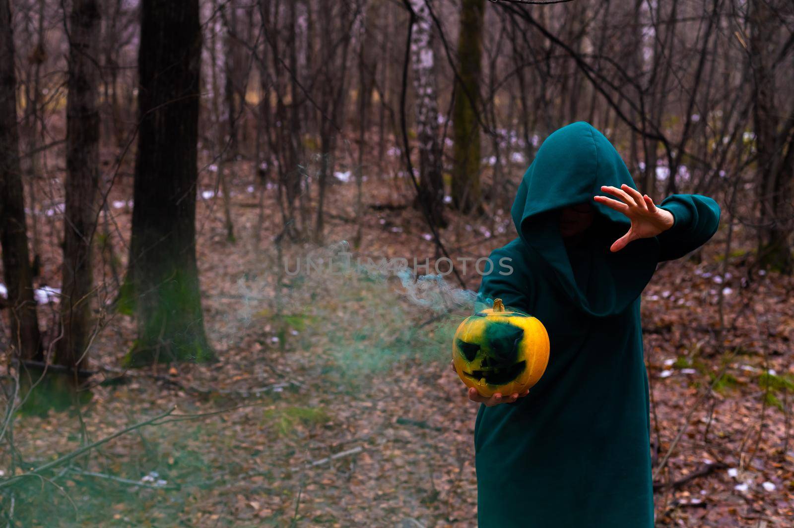 A creepy witch holds a steaming pumpkin in a deep forest. Jack o lantern with green smoke for halloween.