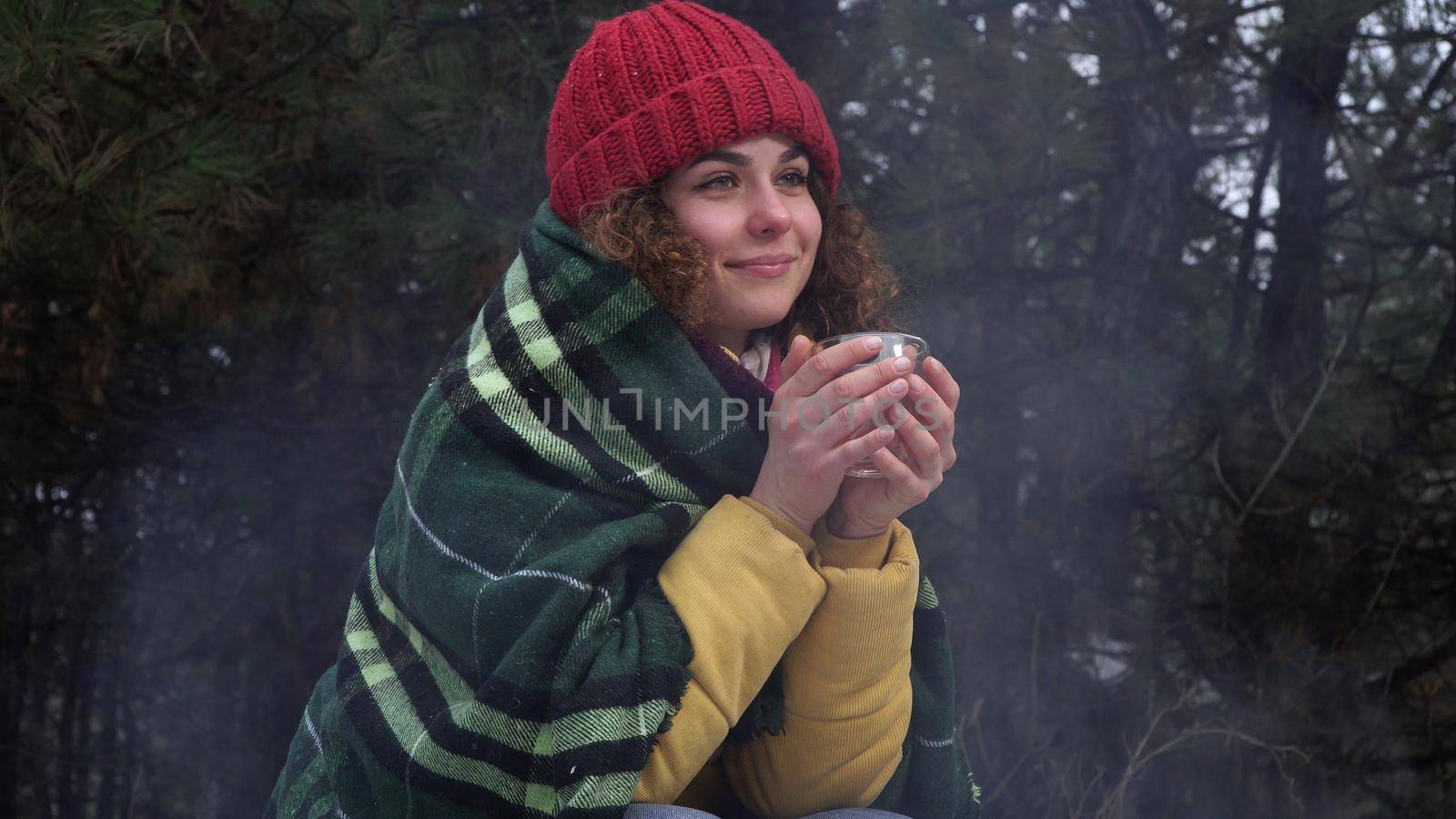 Young woman drinks tea by the fire in the winter forest. She is smiling and happy. Travel and adventure concept.