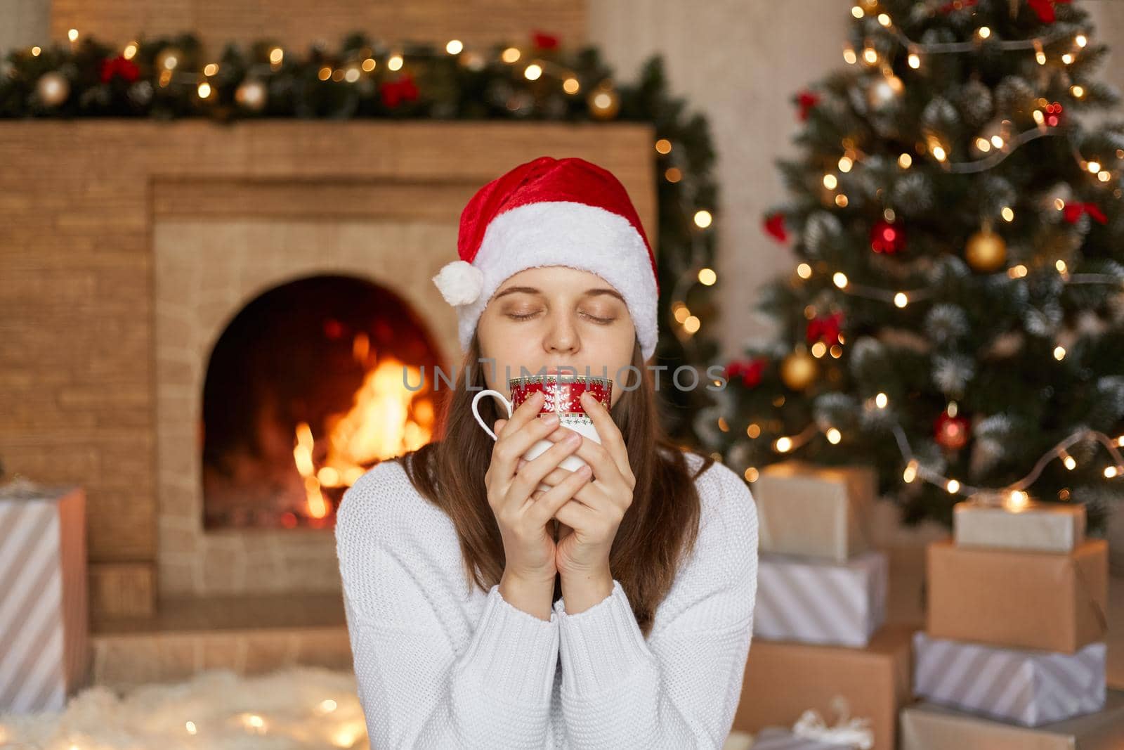 Christmas woman drinking hot tea or coffee, enjoys beverage with closed eyes, holding cup with both hands, sitting in living room with Christmas decoration, girl in warm sweater and santa hat.