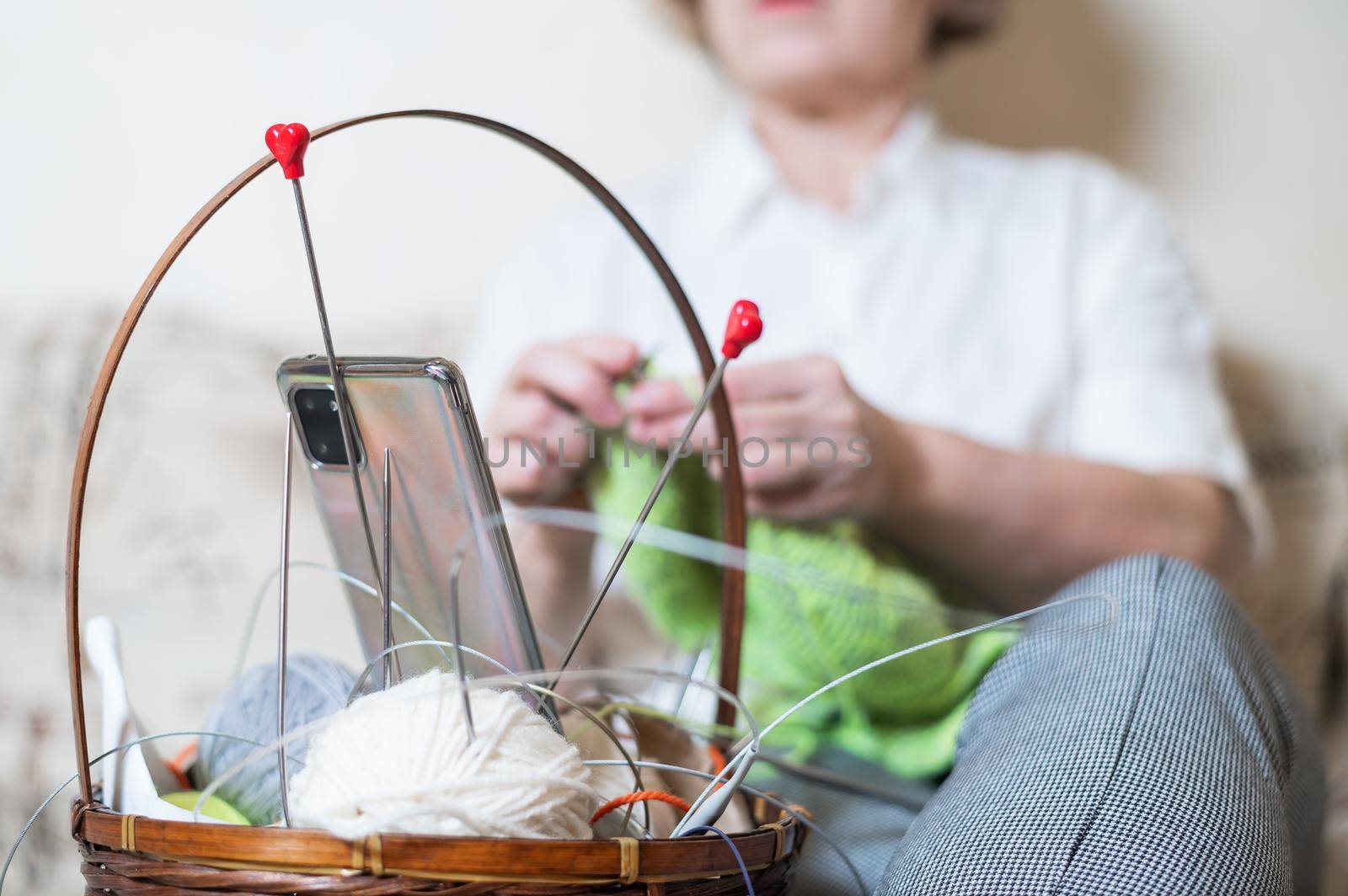 Faceless old woman knits and watches online training on a smartphone. Close-up of female hands with yarn and knitting needles. by mrwed54