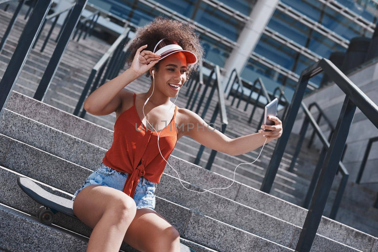 Young woman wearing tennis visor and earphones free style on the street sitting on concrete stairs on skateboard taking selfie photos on smartphone smiling playful