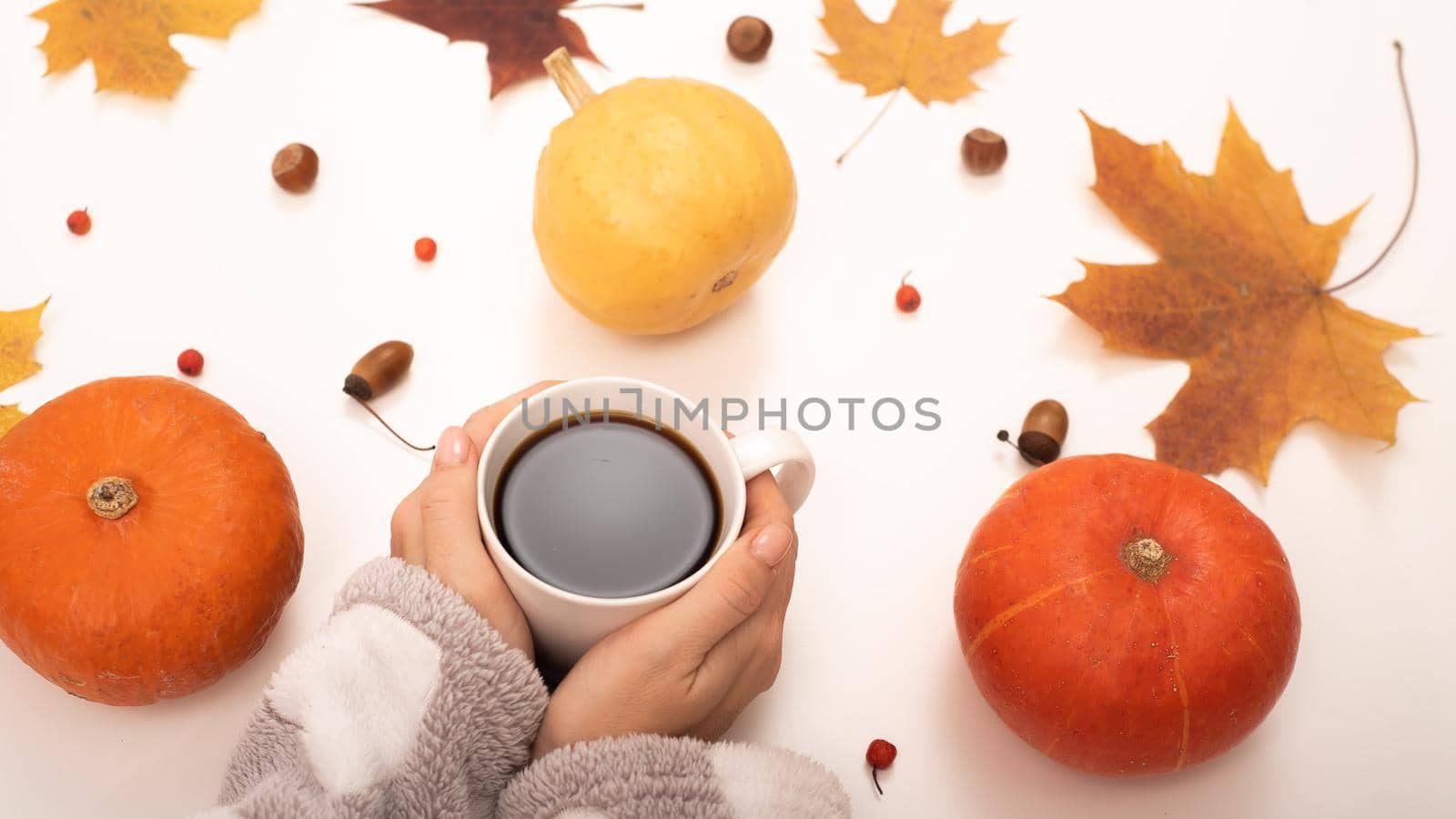 A woman holds a cup of black coffee near the yellow maple leaves of a pumpkin on a white background. Autumn flat lay