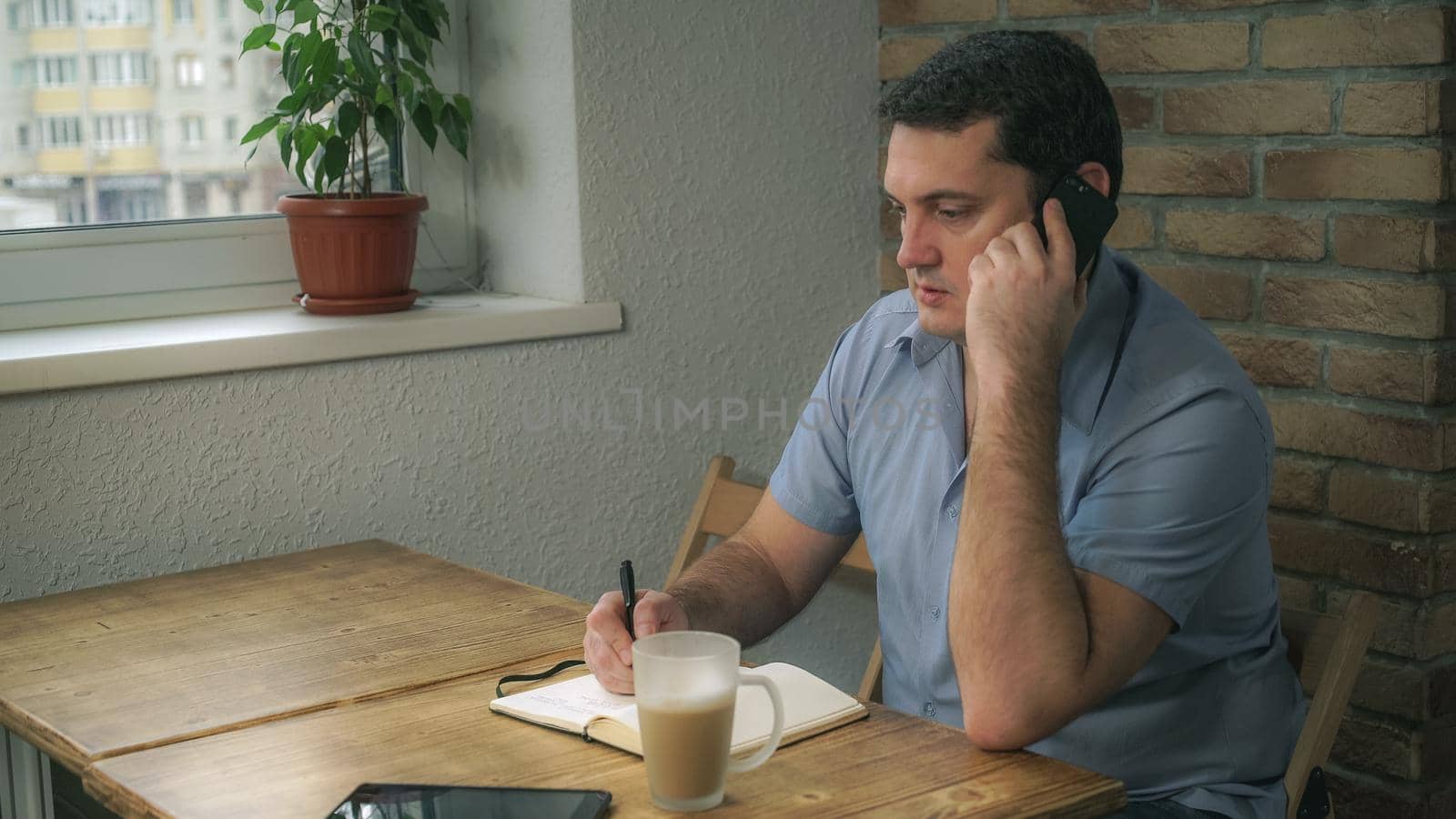 Attractive adult man resting in a cafe and talks on the phone, taking notes in a diary, is a cup of coffee next to lay tablet on the table.