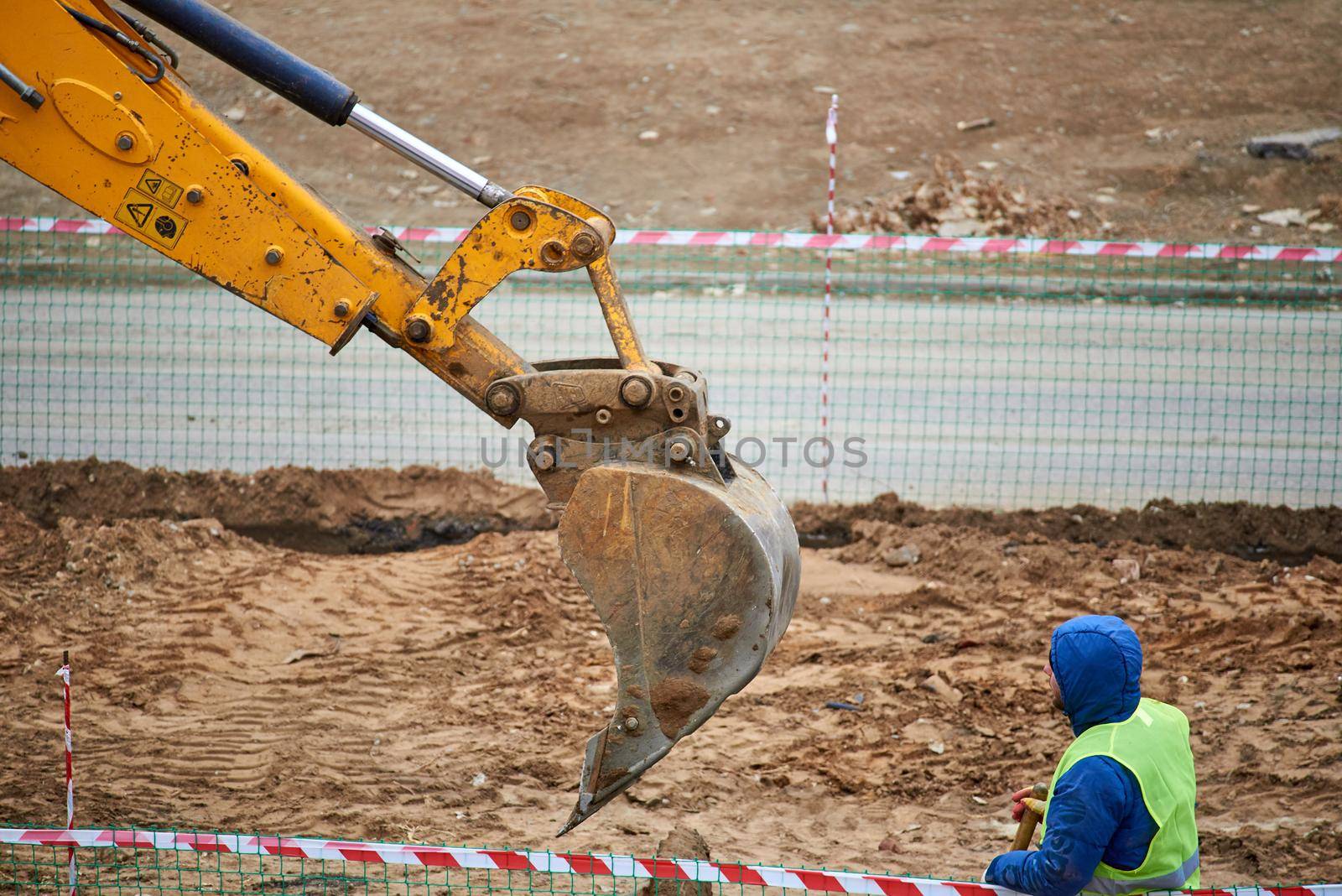 Astrakhan, Russia - 02.15.2021: A worker in a blue jacket and green signal vest he watches the backhoe bucket dig into the ground