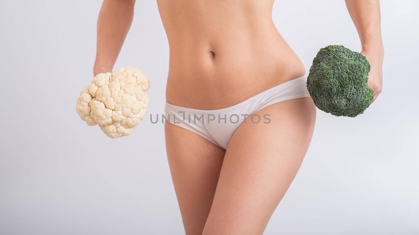 A faceless woman in panties holds cauliflower and broccoli on a white background. Food habits. by mrwed54
