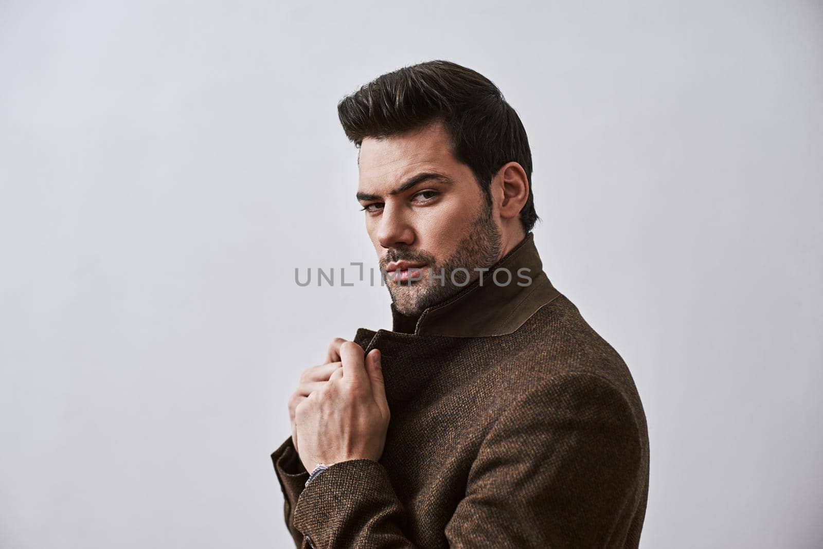 Isolated studio shot of handsome young male worker with bristle and stylish haircut holding his brown jacket while standing over white background and looking at camera.