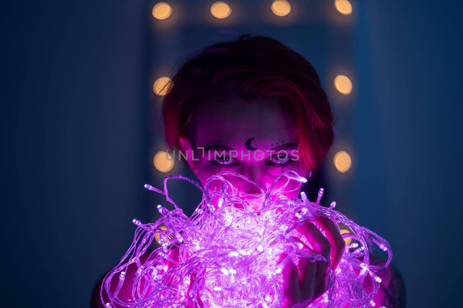 Woman with witch makeup and an earring in her nose. The girl holds a garland of flickering pink lights near her face. Light bulbs illuminate the witch's face in the dark. by mrwed54