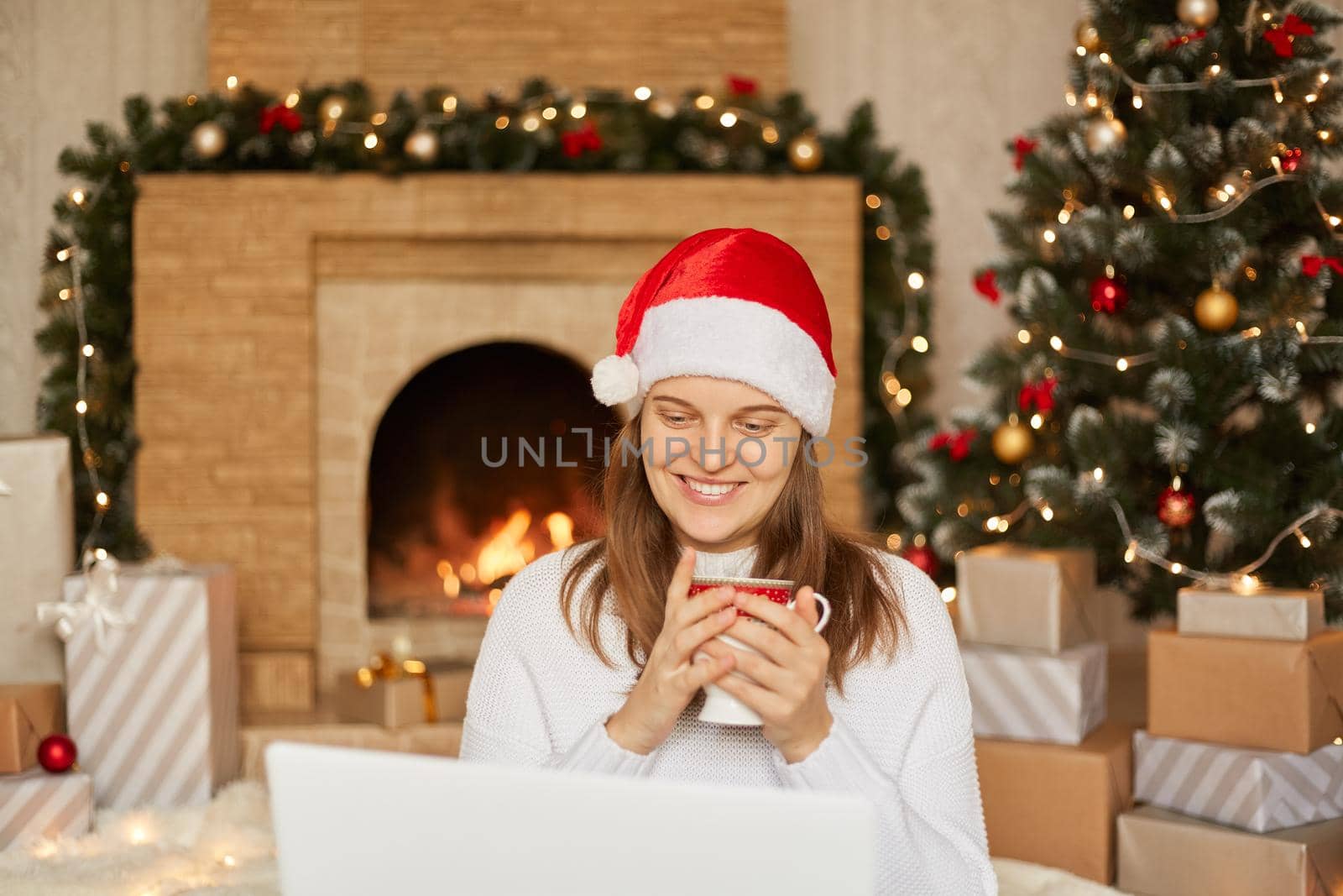Photo of young lady sit in front of laptop, holding cup with hot tea, look at screen screen, wearing red santa hat and white sweater in decorated x-mas living room, congratulates somebody with holiday