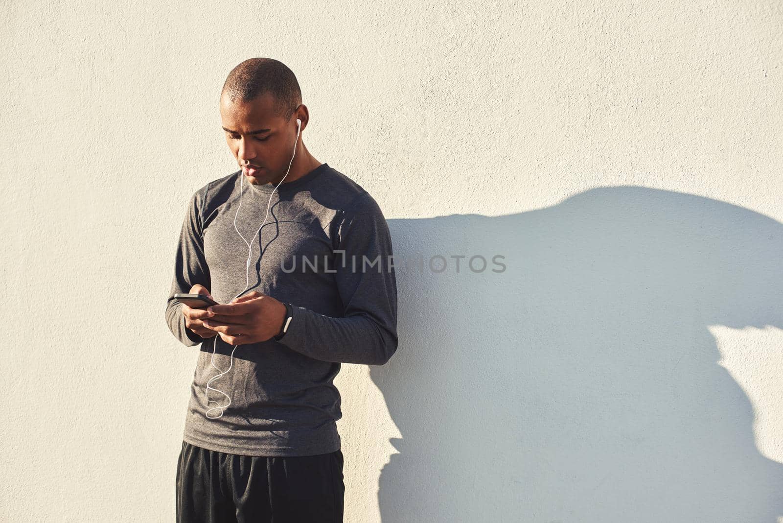 Little break. Close up portrait of motivated africanathlete taking a break and listening music on earphones while standing against the white wall. Cardio training. Morning workout. Sport motivation concept. Fitness