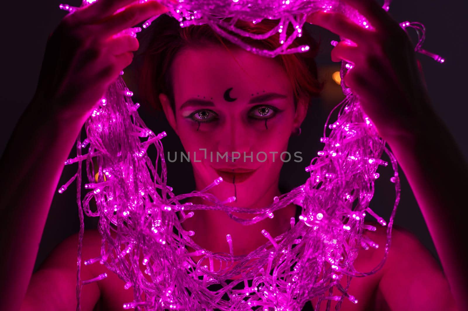 Woman with witch makeup and an earring in her nose. The girl holds a garland of flickering pink lights near her face. Light bulbs illuminate the witch's face in the dark. by mrwed54