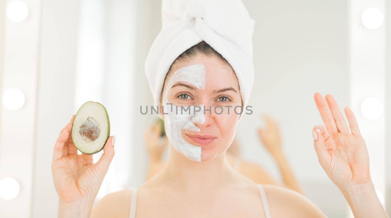A woman with a towel on her hair and in a clay face mask holds an avocado. Taking care of beauty at home.
