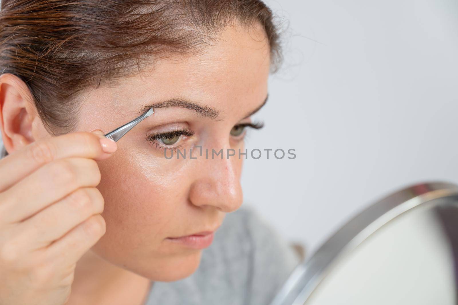 Caucasian woman looks in the mirror and does the eyebrow correction herself with tweezers by mrwed54