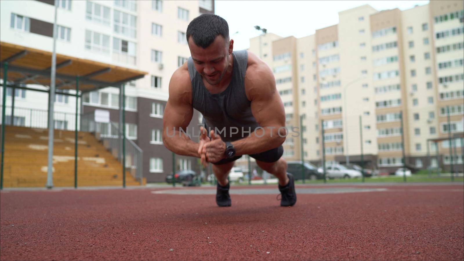 Young muscular man doing push up exercise on the ground outdoors. He looking at camera. The guy presses on the sports ground on the street.