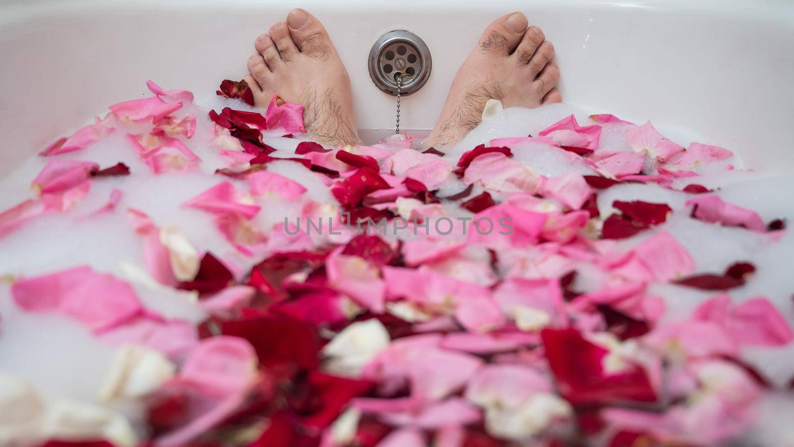 Funny picture of a man taking a relaxing bath. Close-up of male feet in a bath with foam and rose petals.