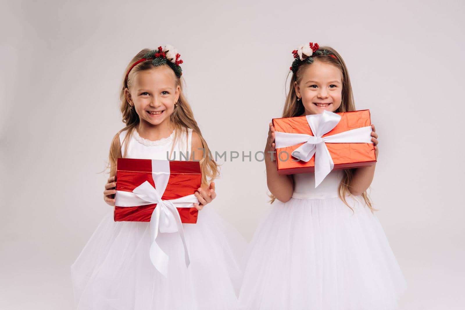 two little girls in white dresses with Christmas gifts on a white background smile.