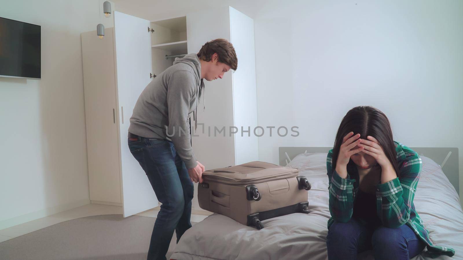 Husband and wife quarreled. Young man packing clothes in suitcase. Upset woman sitting on the bed. Girlfriend and boyfriend at home. Flat with casual interior.