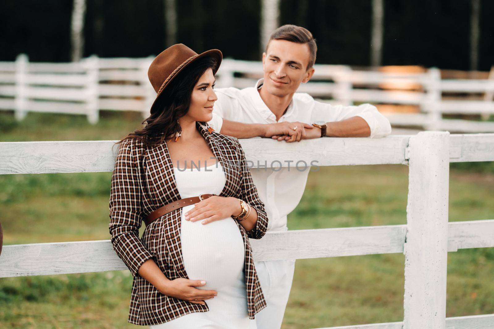 a pregnant girl in a hat and her husband in white clothes stand next to a horse corral at sunset.a stylish couple is waiting for a child in nature.