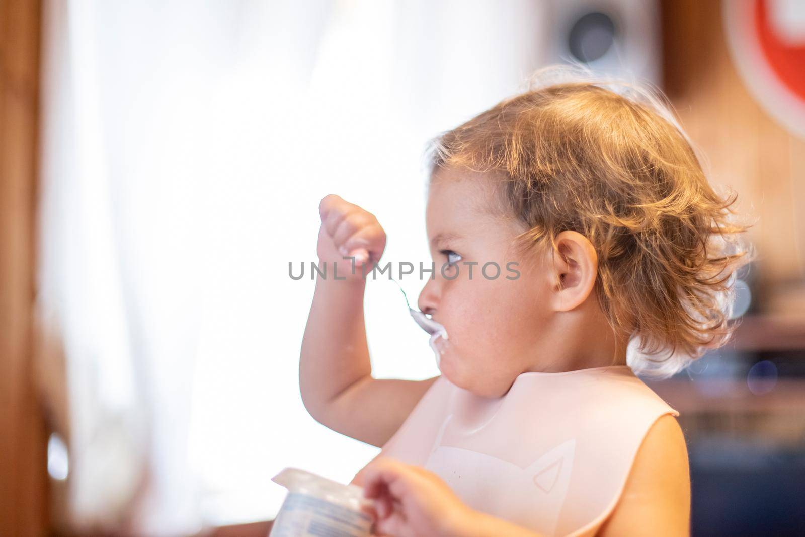 cute baby eating yogurt with spoon by himself. adorable toddler has lunch. hungry child eas food