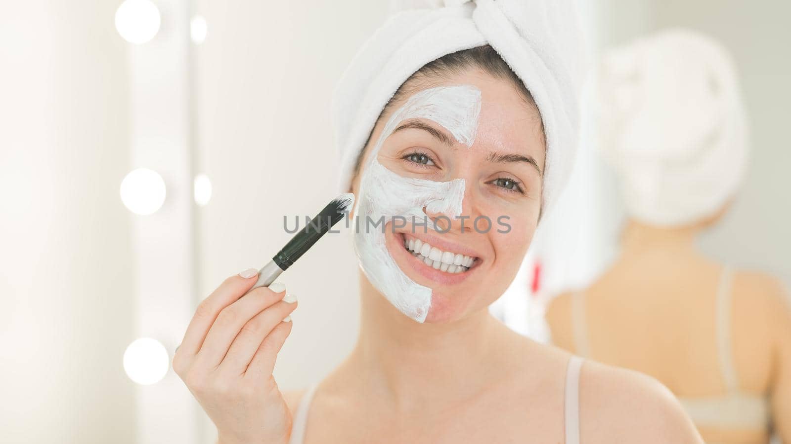 Beautiful caucasian woman with a towel on her hair applies a clay face mask. Taking care of beauty at home.