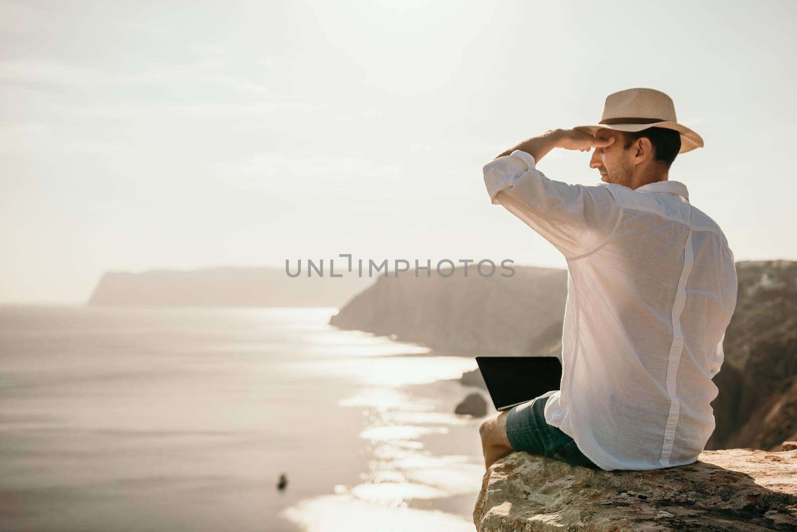 Digital nomad, man in the hat, a businessman with a laptop sits on the rocks by the sea during sunset, makes a business transaction online from a distance. Remote work on vacation. by panophotograph