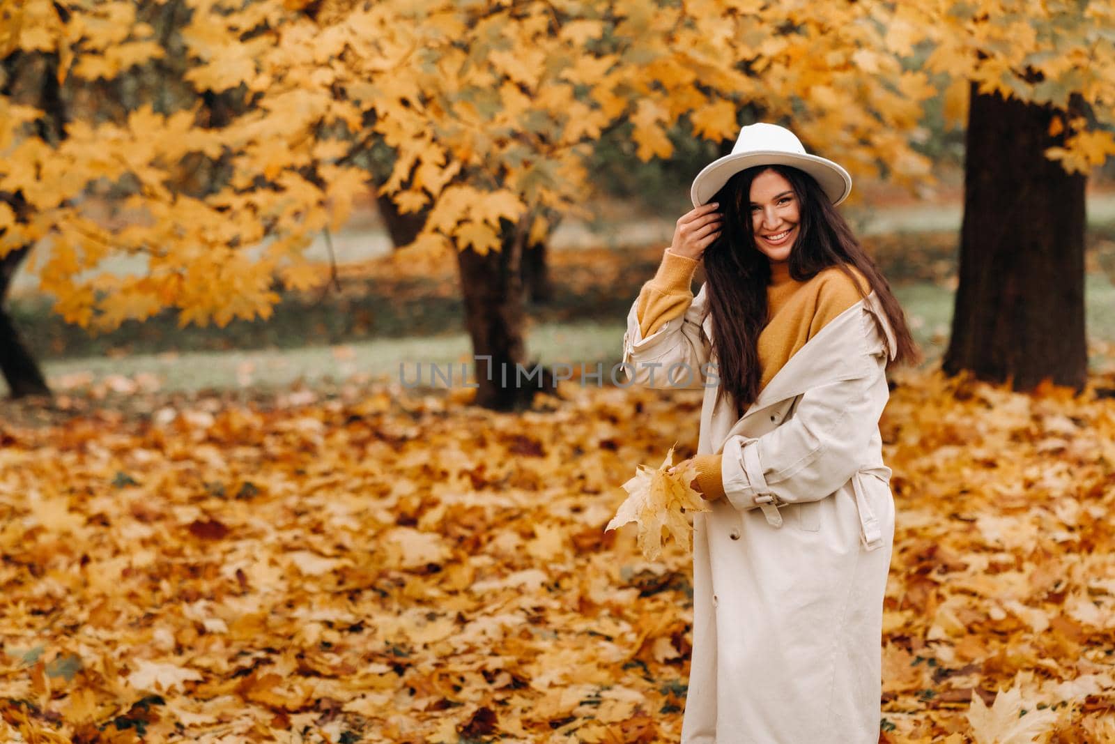 a cheerful girl in a white coat and hat smiles in an autumn Park.portrait of a smiling woman in Golden autumn. by Lobachad