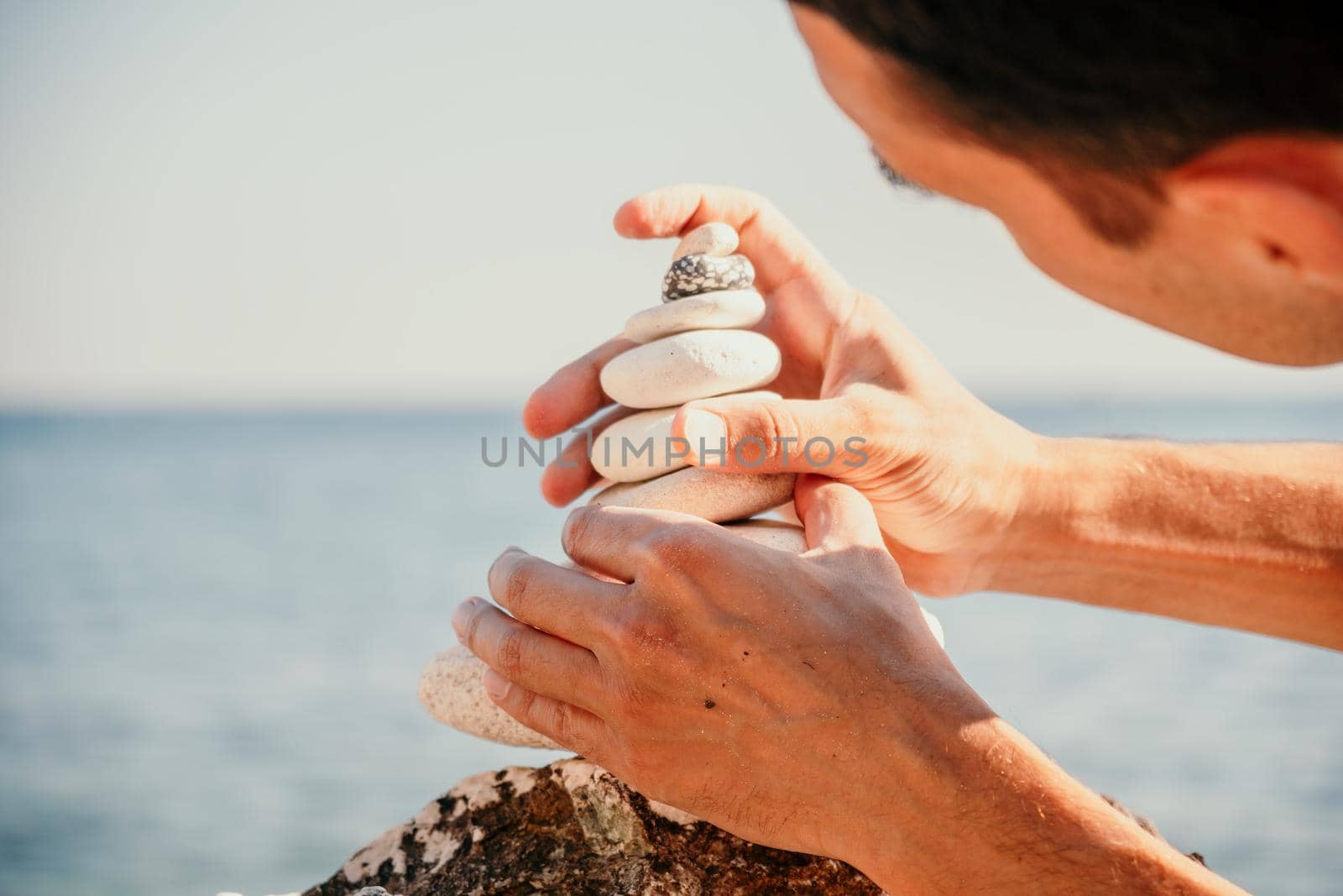 Man bilds stones pyramid on the seashore on a sunny day on the blue sea background. Happy holidays. Pebble beach, calm sea, travel destination. Concept of happy vacation on the sea, meditation, spa by panophotograph