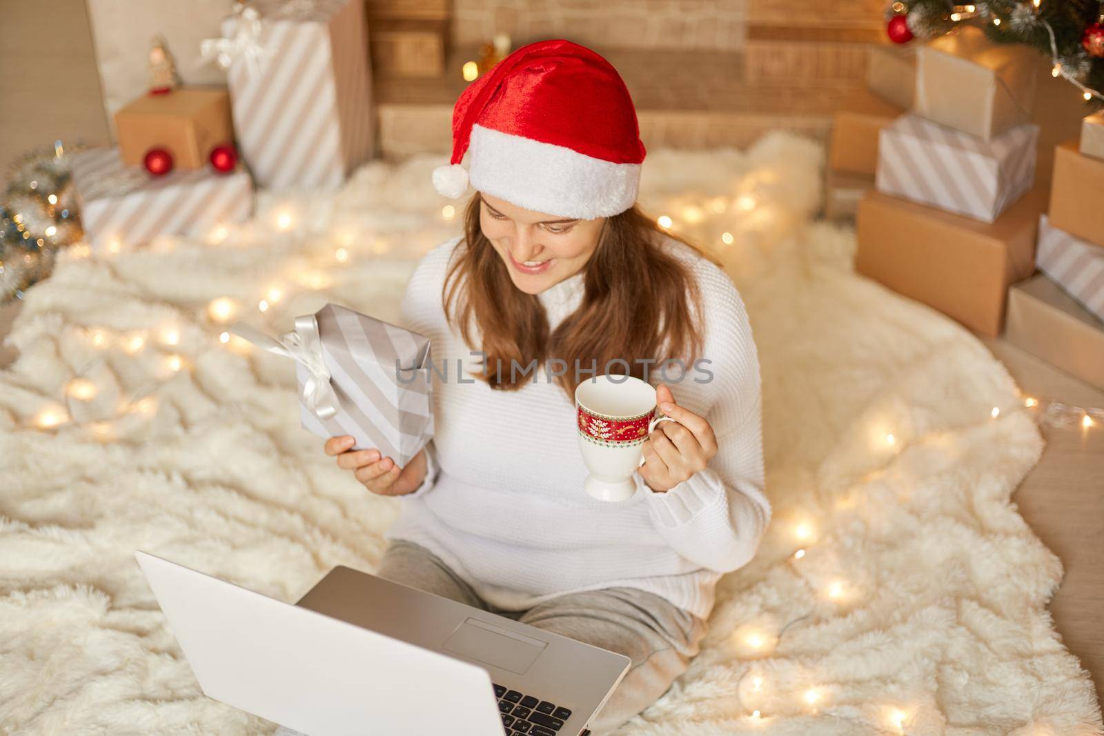 Lovely female having video call with their family and showing Christmas present to camera, woman in santa hat and white sweater with giftbox using laptop with web cam for greeting close people.. by sementsovalesia