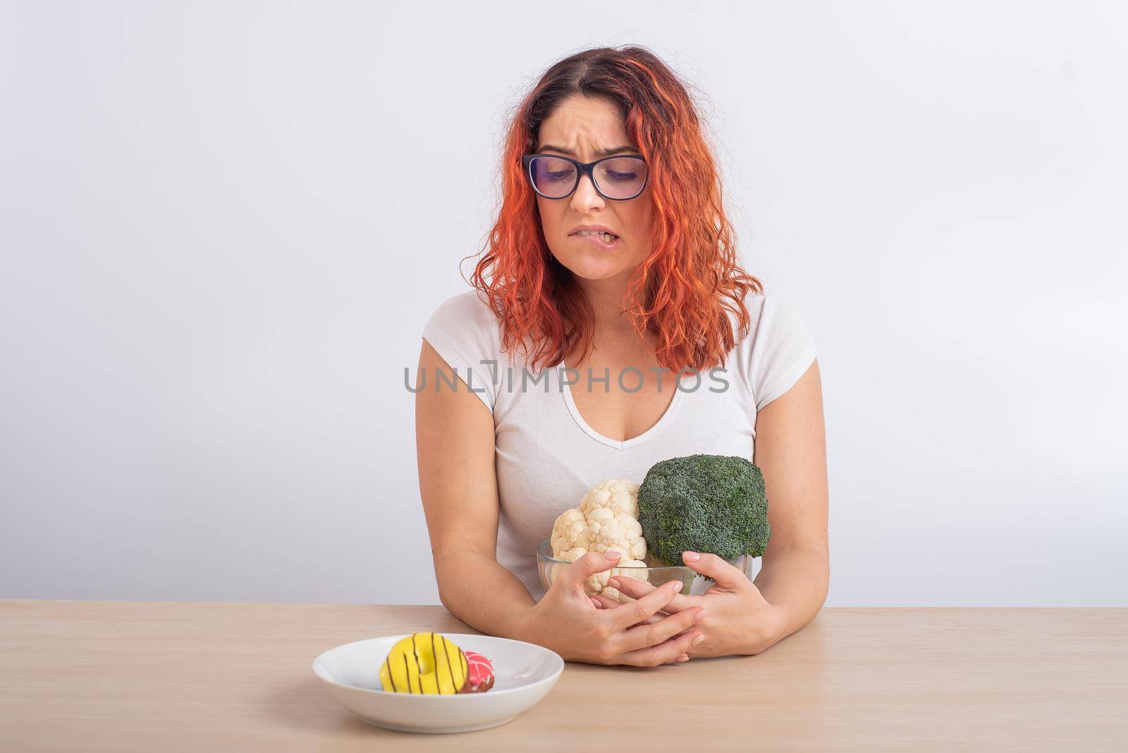 Caucasian woman on a diet dreaming of fast food. Redhead girl chooses between broccoli and donuts on white background. by mrwed54