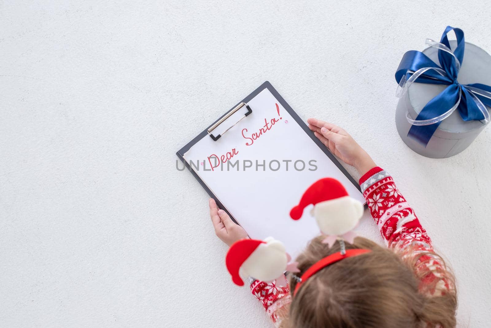 Child writing letter to Santa Claus at Christmas on white background. Top view. notepad for wishes, gift list.Girl in red dress lying and writing christmas letter.celebration concept. by YuliaYaspe1979