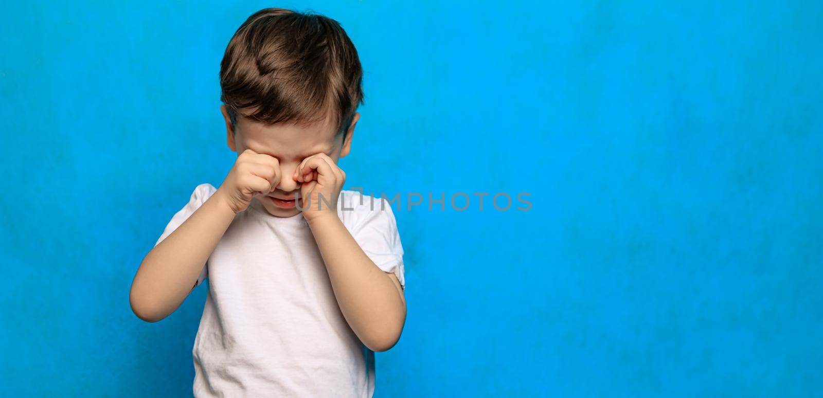 A boy on a blue background rubs his eyes . Eye health. Eye diseases. A crying baby. Fatigue of children. Psychology. Copy Space