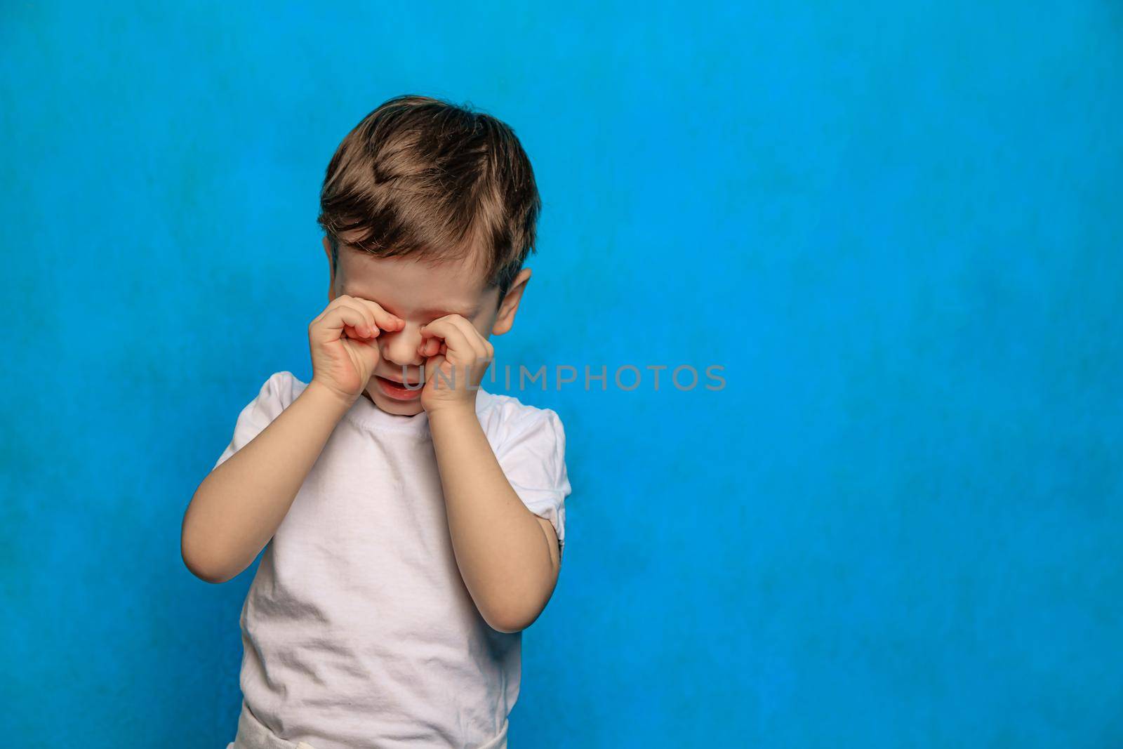A boy on a blue background rubs his eyes . Eye health. Eye diseases. A crying baby. Fatigue of children. Psychology. Copy Space