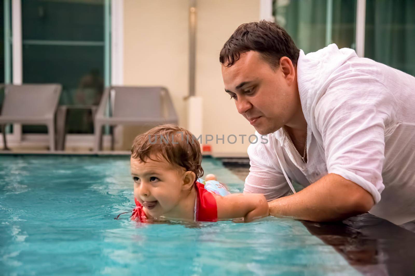 father teaches cute toddler little girl in swimsuit to swim in the pool holding his hand