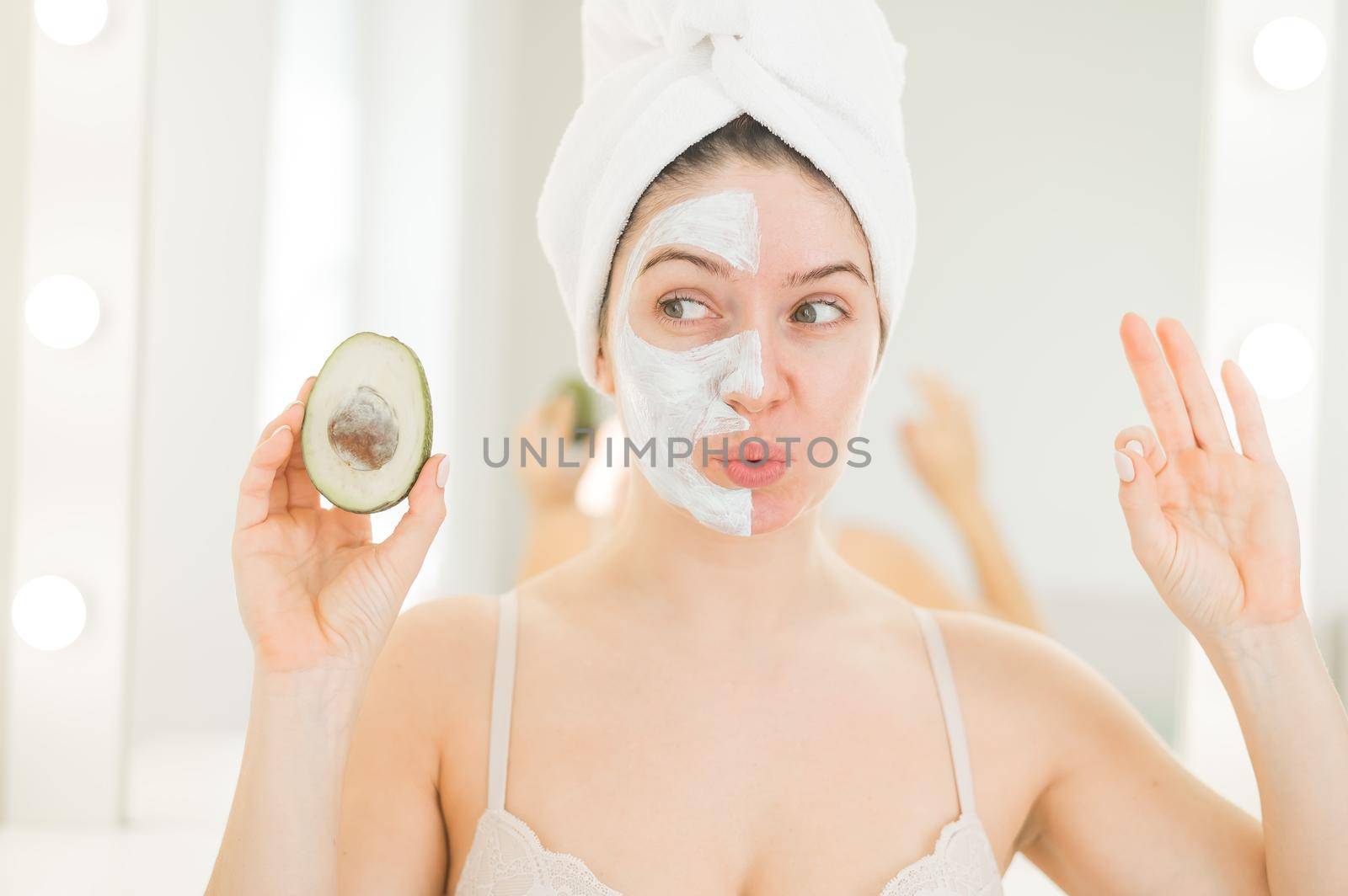 A woman with a towel on her hair and in a clay face mask holds an avocado. Taking care of beauty at home by mrwed54