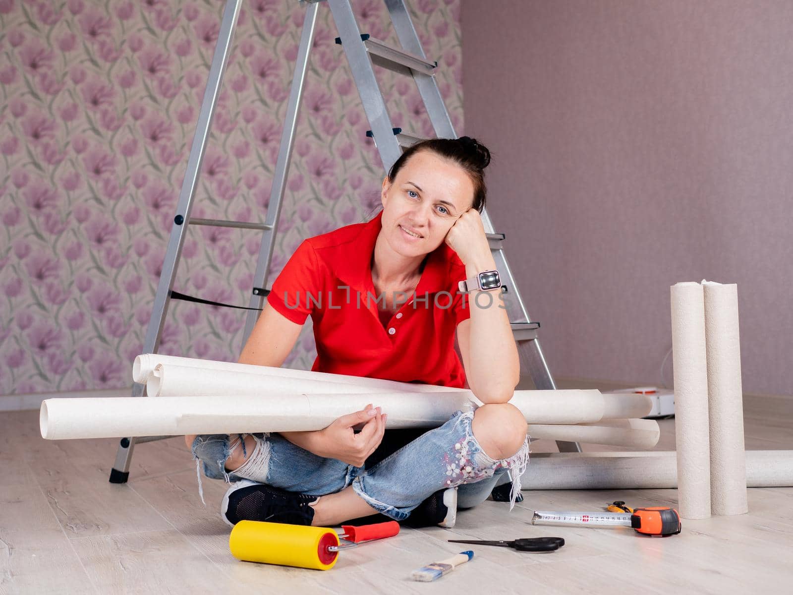 Portrait of a woman sitting on the floor with rolls of Wallpaper in her hands on the background of a stepladder.