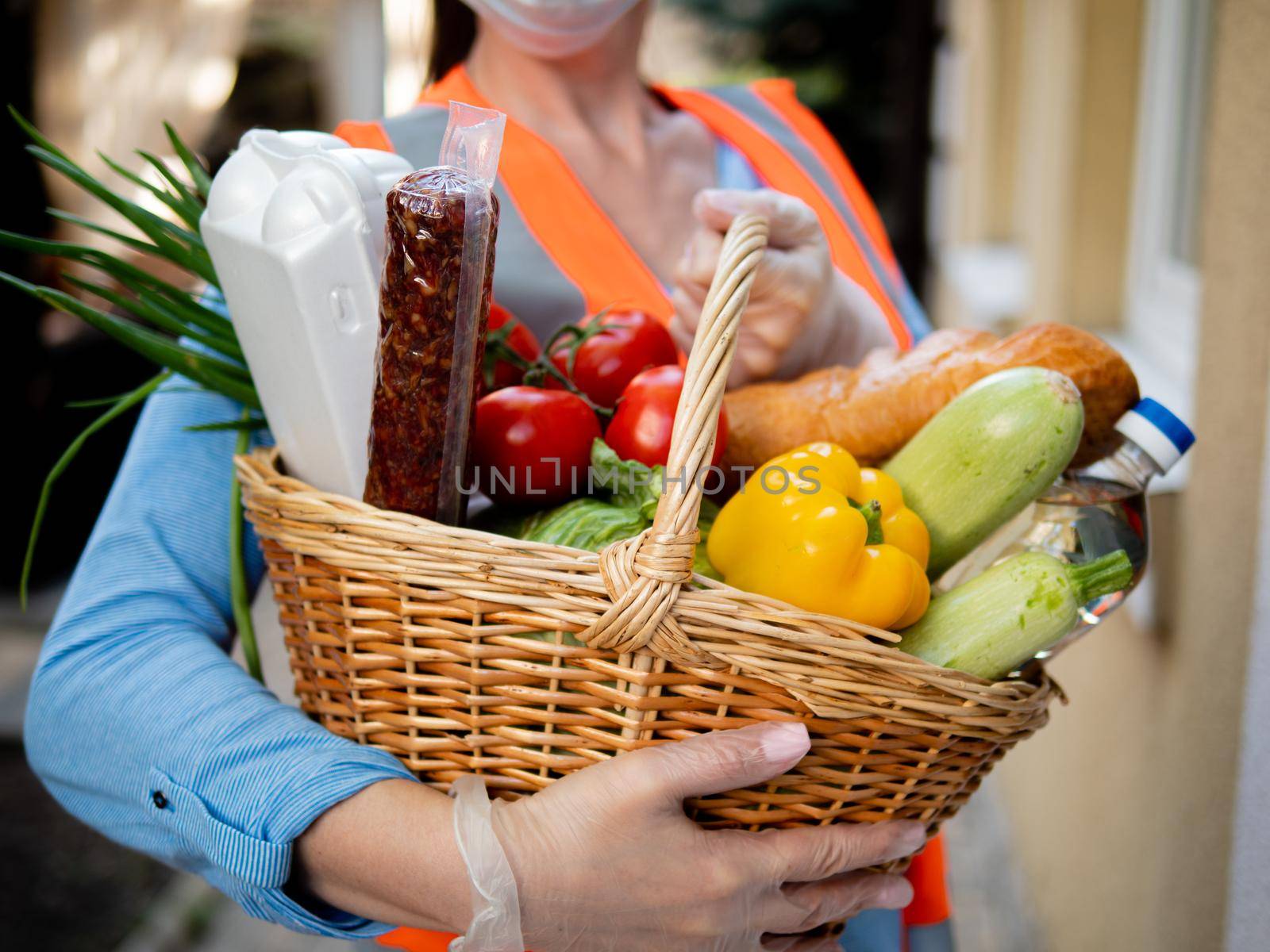 A gloved woman holds a wicker basket full of vegetables and other essential food items. Volunteers deliver food packages during the period of the pandemic. by Utlanov